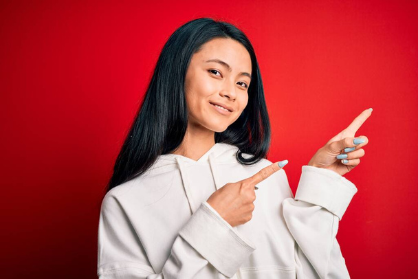 Genç ve güzel Çinli sporcu kadın izole edilmiş kırmızı arka plan üzerinde sweatshirt giyiyor gülümsüyor ve kameraya bakıyor iki eliyle ve parmaklarıyla kenarı işaret ediyor.. - Fotoğraf, Görsel