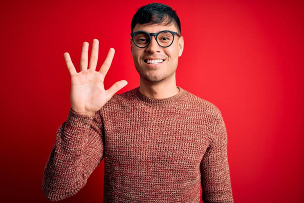 Jovem hispânico bonito vestindo óculos nerd sobre fundo vermelho mostrando e apontando para cima com os dedos número cinco enquanto sorri confiante e feliz
. - Foto, Imagem