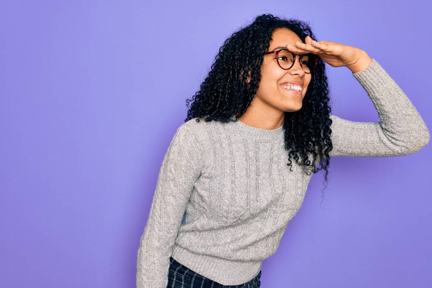 Jeune femme afro-américaine portant un pull décontracté et des lunettes sur fond violet très heureuse et souriante regardant loin avec la main au-dessus de la tête. Concept de recherche
. - Photo, image