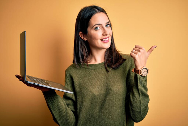 Junge brünette Frau mit blauen Augen, die mit Computer-Laptop über gelbem Hintergrund arbeitet Zeigt nach hinten mit Hand und Daumen nach oben, lächelt selbstbewusst - Foto, Bild