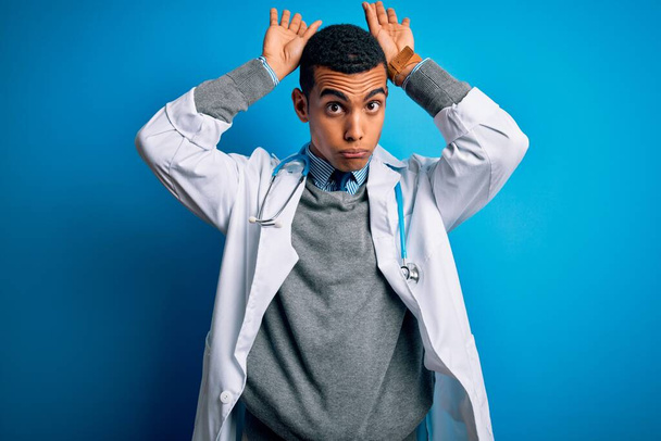 Bello afroamericano medico uomo indossando cappotto e stetoscopio su sfondo blu Fare coniglietto orecchie gesto con le mani palme cercando cinico e scettico. Concetto di coniglio pasquale
. - Foto, immagini