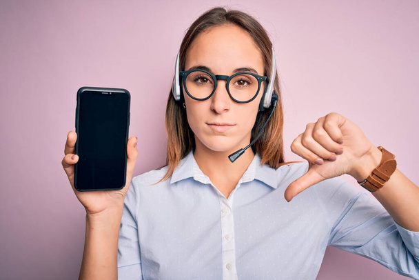 Beautiful call center agent woman working using headset holding smartphone holding screen with angry face, negative sign showing dislike with thumbs down, rejection concept - Foto, afbeelding
