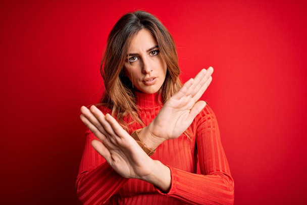 Young beautiful brunette woman wearing casual turtleneck sweater over red background Rejection expression crossing arms doing negative sign, angry face - Foto, Bild
