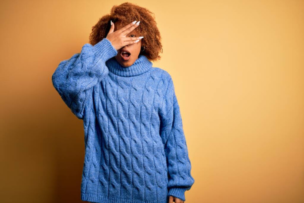 Young beautiful African American afro woman with curly hair wearing blue turtleneck sweater peeking in shock covering face and eyes with hand, looking through fingers with embarrassed expression. - Fotoğraf, Görsel