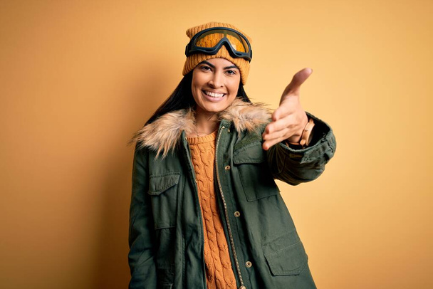 Mladá krásná hispánská žena v lyžařských brýlích a kabátě pro zimní počasí s přátelským úsměvem nabízí potřesení rukou jako pozdrav a přivítání. Úspěšná práce. - Fotografie, Obrázek