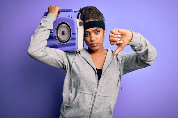 Mladý africký americký afro dívka poslech hudby držení vintage rádio přes fialové pozadí s rozzlobený obličej, negativní znamení ukazující odpor s palci dolů, odmítavý koncept - Fotografie, Obrázek