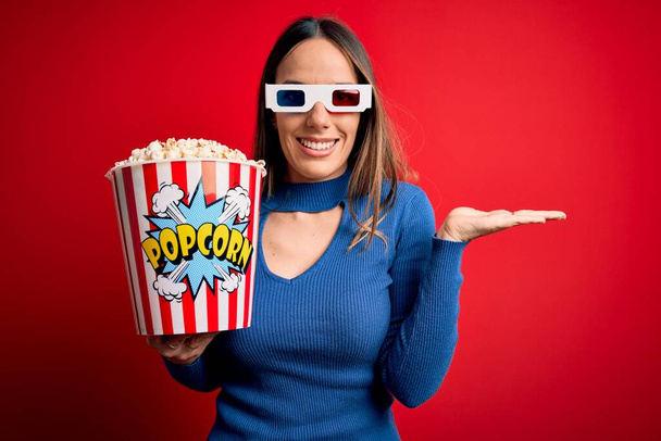 Νεαρή ξανθιά γυναίκα που φοράει 3d γυαλιά και τρώει ποπ κορν βλέποντας μια ταινία στον κινηματογράφο χαμογελώντας χαρούμενα παρουσιάζοντας και δείχνοντας με το χέρι κοιτάζοντας την κάμερα. - Φωτογραφία, εικόνα