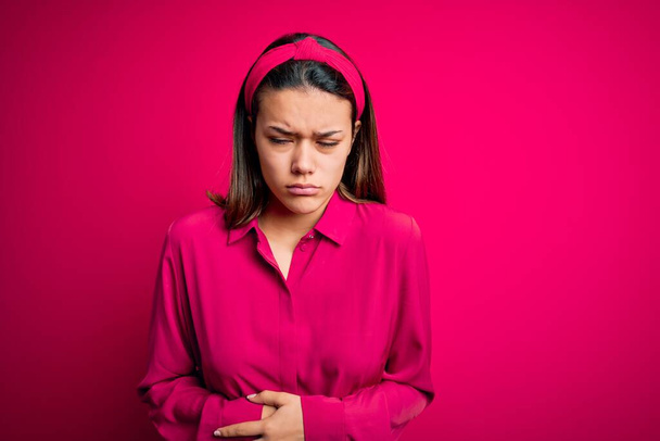 Young beautiful brunette girl wearing casual shirt standing over isolated pink background with hand on stomach because indigestion, painful illness feeling unwell. Ache concept. - Zdjęcie, obraz