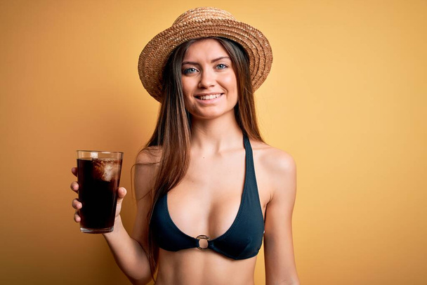 Mladá krásná žena s modrýma očima na dovolené na sobě bikiny a klobouk pití cola nápoj s veselou tváří stojí a usmívá se s sebevědomým úsměvem ukazující zuby - Fotografie, Obrázek