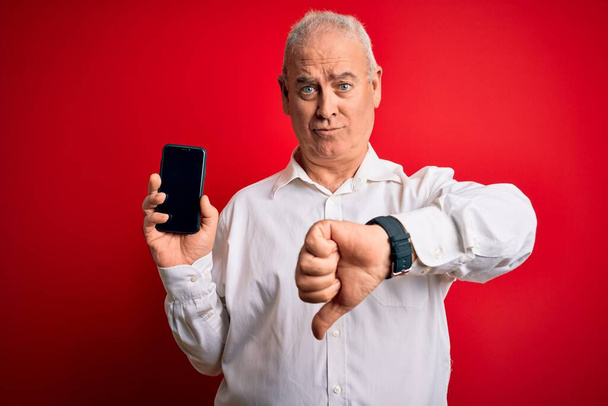 Mezzo età hoary uomo tenendo smartphone mostrando schermo su sfondo rosso isolato con faccia arrabbiata, segno negativo mostrando antipatia con i pollici verso il basso, concetto di rifiuto
 - Foto, immagini