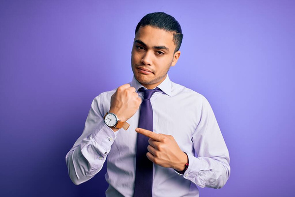 Nuori brasilialainen liikemies yllään tyylikäs solmio seisoo eristetty violetti tausta kiire osoittaa katsella aikaa, kärsimättömyys, katsot kameraa rento ilme
 - Valokuva, kuva