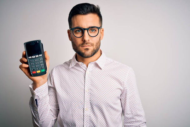 Young handsome man doing payment using dataphone over isolated white background with a confident expression on smart face thinking serious - Photo, Image