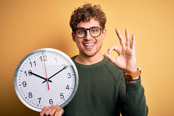 Mladý blonďatý muž s kudrnatými vlasy nosí brýle drží velké hodiny přes žluté pozadí dělá ok znamení s prsty, vynikající symbol - Fotografie, Obrázek