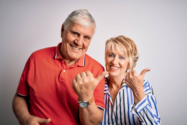 Senior beautiful couple standing together over isolated white background Pointing to the back behind with hand and thumbs up, smiling confident - Photo, image