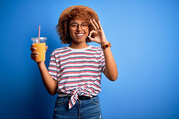 Young African American woman with curly hair on vacation wearing hat drinking orange juice doing ok sign with fingers, excellent symbol - Fotografie, Obrázek