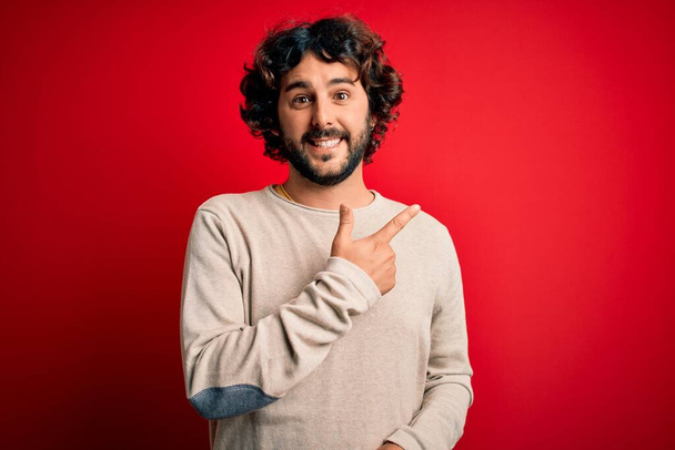 Young handsome man with beard wearing casual sweater standing over red background cheerful with a smile of face pointing with hand and finger up to the side with happy and natural expression on face - Photo, Image