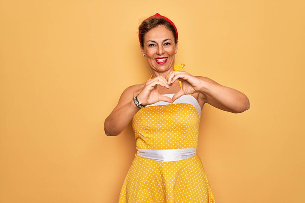 手でハートシンボル形状を行う愛に笑みを浮かべて黄色の背景に50スタイルのレトロなドレスを身に着けている女性をピンアップ中世のシニア。ロマン主義. - 写真・画像