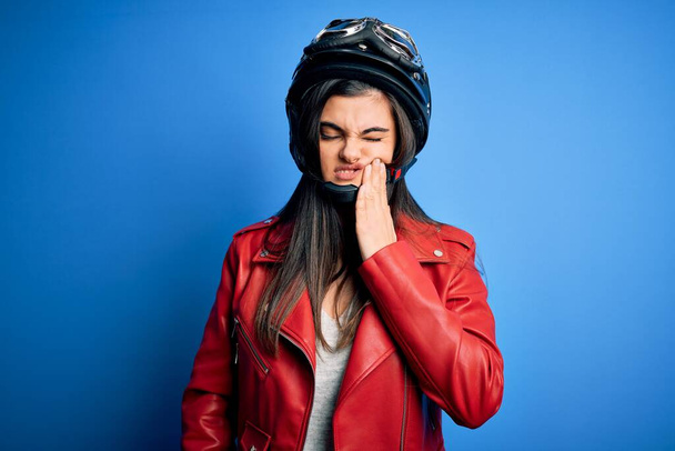 歯の歯痛や歯の病気のために痛みを伴う式で手で口に触れるオートバイのヘルメットとジャケットを身に着けている若い美しいブルネットのオートバイの女性。歯医者 - 写真・画像