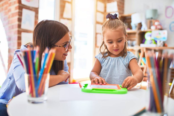 白人の女の子の子供は女教師と遊び場で遊んで学びます.母と娘-おもちゃの周りにプレイルームで磁気黒板に描く - 写真・画像