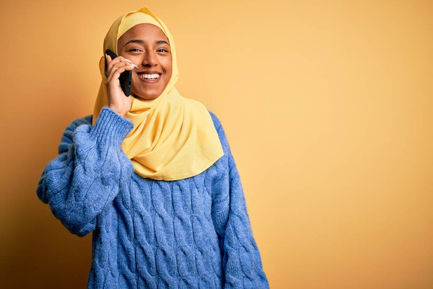 Афроамериканка в мусульманском хиджабе разговаривает по смартфону со счастливым лицом стоя и улыбаясь с уверенной улыбкой, показывая зубы
 - Фото, изображение