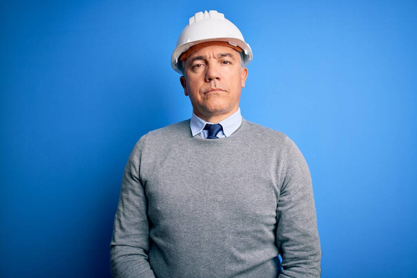 Middle age handsome grey-haired engineer man wearing safety helmet over blue background Relaxed with serious expression on face. Simple and natural looking at the camera. - Photo, Image