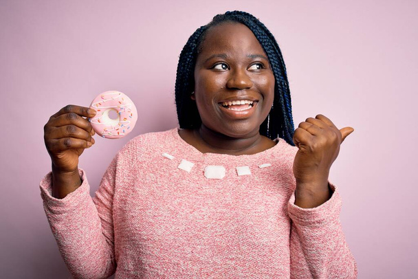 Mladý africký Američan plus velikost žena s copánky jíst koblihu přes růžové pozadí ukazuje palcem nahoru na stranu se šťastnou tváří usmívá - Fotografie, Obrázek