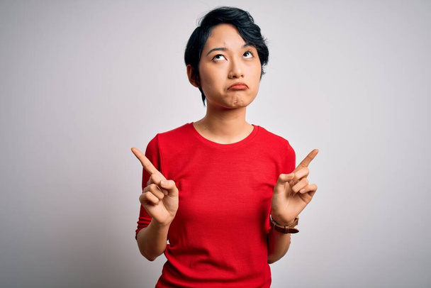 Mladá krásná asijská dívka v ležérní červené tričko stojící nad izolovaným bílým pozadím ukazující nahoru vypadá smutně a rozrušeně, indikující směr s prsty, nešťastné a depresivní. - Fotografie, Obrázek