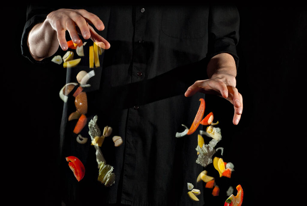 A man in a black shirt prepares food on a dark background. Meat, potatoes, tomatoes, carrots and other vegetables fly in the air. Blurred as an artistic effect. Motion, flight and levitation concept. - Valokuva, kuva