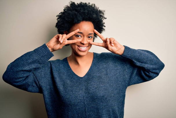 jong mooi Afro-Amerikaans afro vrouw met krullend haar dragen casual trui doet vrede symbool met vingers over het gezicht, glimlachend vrolijk tonen overwinning - Foto, afbeelding
