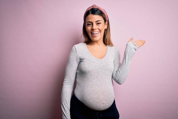 Mladá krásná brunetka žena těhotná čeká dítě přes izolované růžové pozadí s úsměvem veselý prezentaci a ukazuje s dlaní ruky při pohledu na fotoaparát. - Fotografie, Obrázek