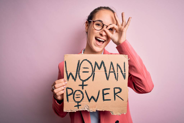 Jovem bela ativista mulher pedindo direitos das mulheres segurando banner com mensagem de poder com o rosto feliz sorrindo fazendo ok sinal com a mão no olho olhando através dos dedos
 - Foto, Imagem