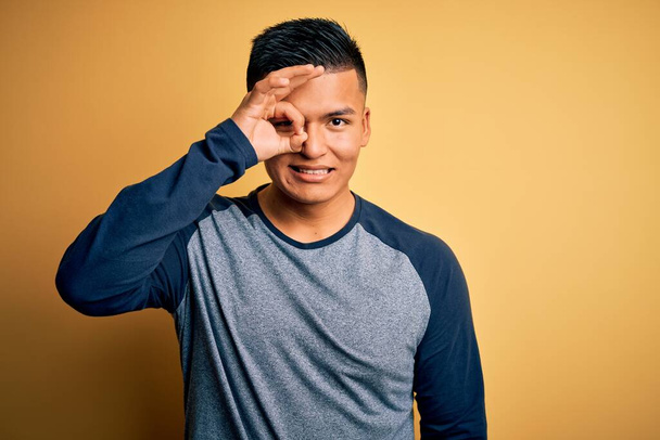 Молодой красивый латиноамериканец в обычной футболке, стоящей на жёлтом фоне, делает хорошо жест с улыбкой на руке, глаза смотрят сквозь пальцы со счастливым лицом
. - Фото, изображение