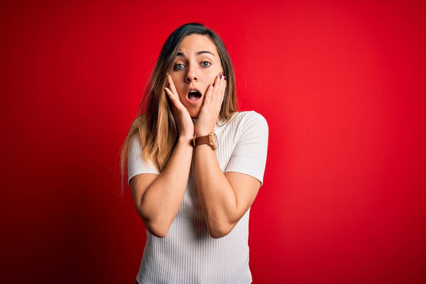 Beautiful blonde woman with blue eyes wearing casual white t-shirt over red background afraid and shocked, surprise and amazed expression with hands on face - Foto, Bild