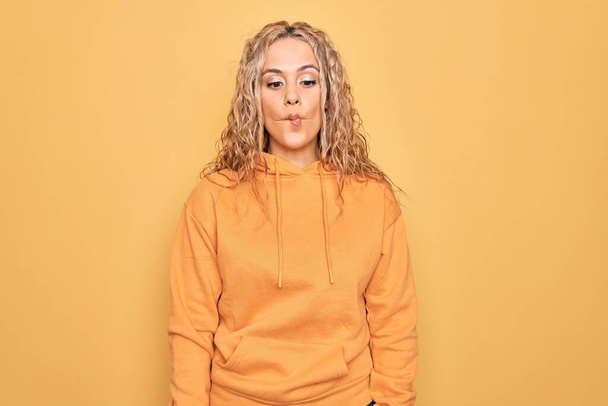 Young beautiful blonde sporty woman wearing casual sweatshirt over yellow background making fish face with lips, crazy and comical gesture. Funny expression. - Photo, Image