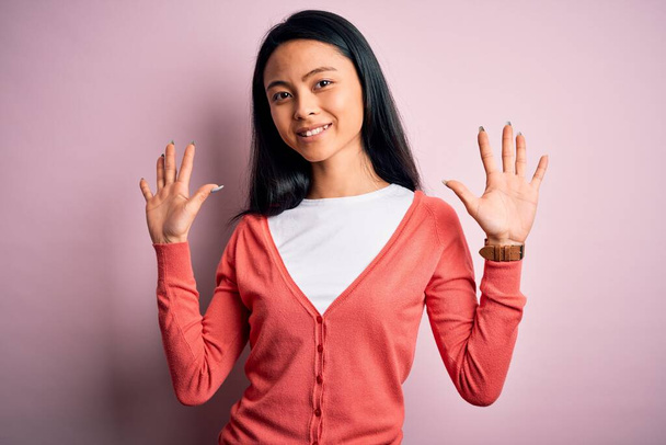 Jovem mulher chinesa bonita vestindo camisola casual sobre fundo rosa isolado mostrando e apontando para cima com os dedos número dez enquanto sorri confiante e feliz
. - Foto, Imagem