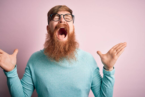 Красивый ирландский рыжий мужчина с бородой в очках на розовом изолированном фоне празднует безумный и безумный успех с поднятыми руками и закрытыми глазами, крича взволнованно. Концепция победителя
 - Фото, изображение
