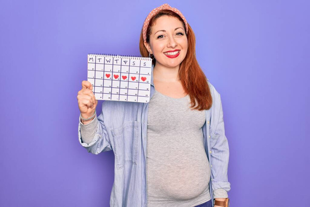 Fiatal gyönyörű vörös hajú terhes nő vár baba gazdaság időszak naptár boldog arc áll, és mosolygós magabiztos mosollyal mutatja fogak - Fotó, kép