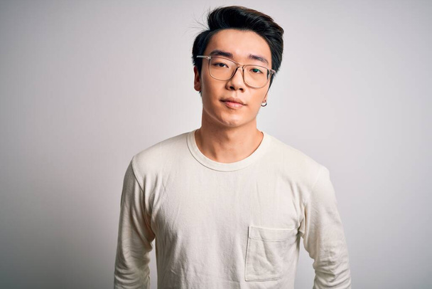 Νεαρός όμορφος κινέζος άντρας που φοράει casual t-shirt και γυαλιά σε λευκό φόντο με σοβαρή έκφραση στο πρόσωπο. Απλή και φυσική κοιτάζοντας την κάμερα. - Φωτογραφία, εικόνα