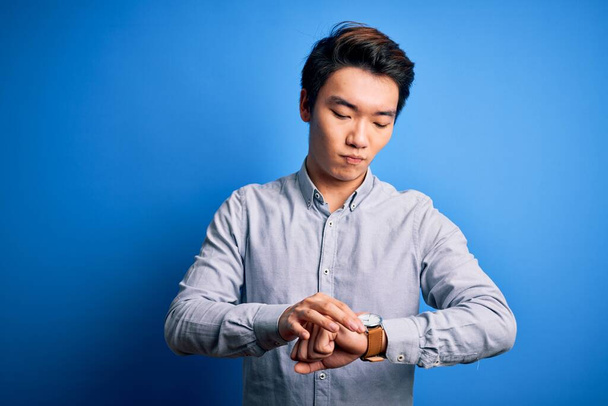 Nuori komea kiinalainen mies päällään rento paita seisoo yli eristetty sininen tausta Tarkistaminen aika rannekellon, rento ja luottavainen
 - Valokuva, kuva