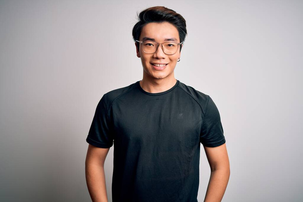 Jeune bel homme chinois portant un t-shirt noir et des lunettes sur fond blanc avec un sourire heureux et cool sur le visage. Personne chanceuse
. - Photo, image