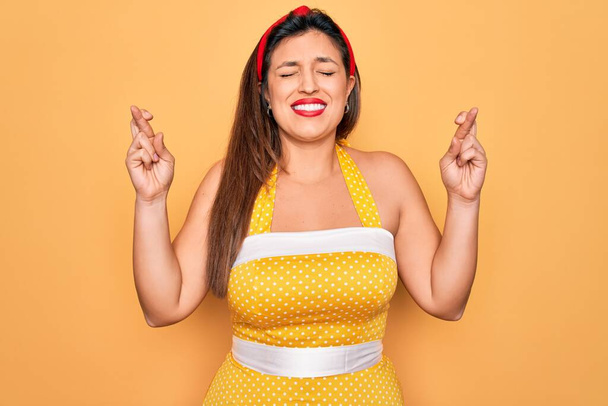 Jovem hispânico pin up mulher vestindo moda sexy estilo dos anos 50 sobre fundo amarelo gesto dedo cruzado sorrindo com esperança e olhos fechados. Sorte e conceito supersticioso
. - Foto, Imagem