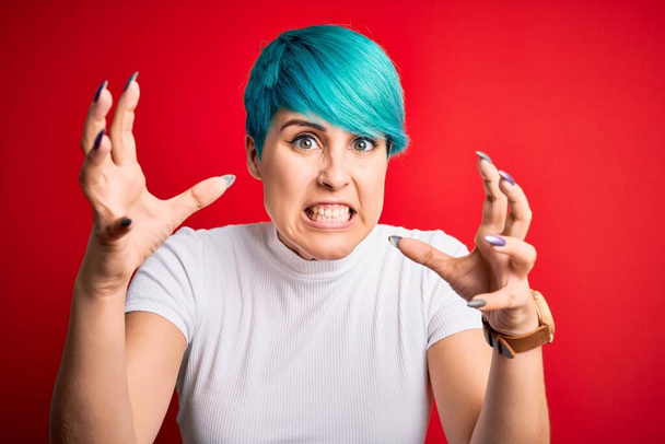 Jovem mulher bonita com cabelo azul moda vestindo t-shirt casual sobre fundo vermelho Grito frustrado com raiva, mãos tentando estrangular, gritando louco
 - Foto, Imagem