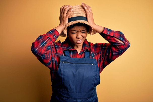 Young African American afro farmer woman with curly hair wearing apron and hat suffering from headache desperate and stressed because pain and migraine. Hands on head. - Fotó, kép