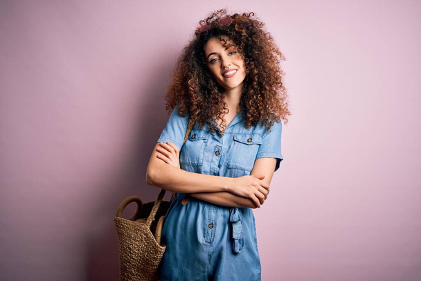 Young beautiful woman with curly hair and piercing wearing denim dress and wicker bag happy face smiling with crossed arms looking at the camera. Positive person. - Photo, image