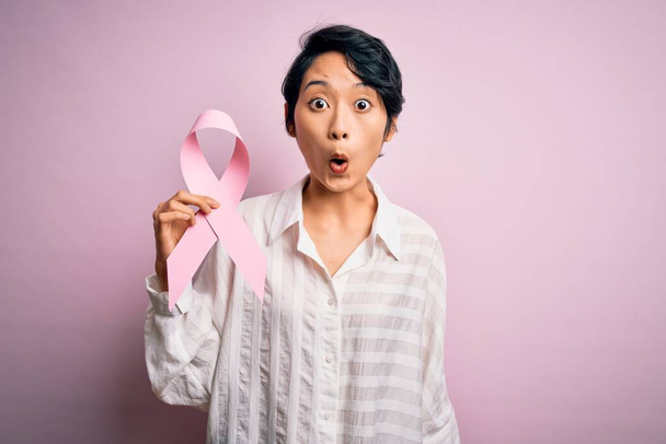 Joven hermosa chica asiática sosteniendo el símbolo de la cinta de cáncer rosa sobre un fondo aislado asustado en shock con una cara sorpresa, asustado y emocionado con la expresión de miedo
 - Foto, Imagen