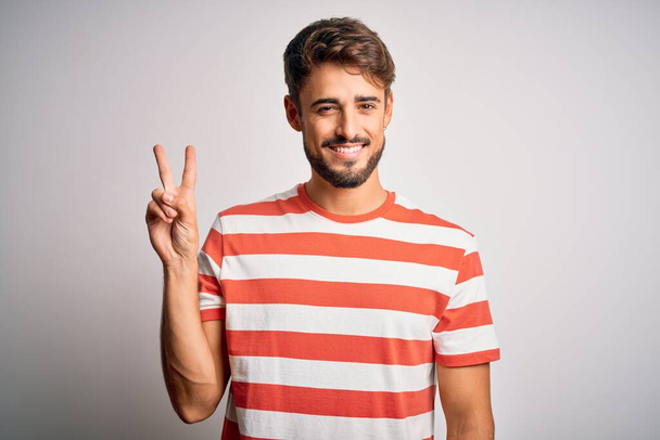 Молодий красивий чоловік з бородою в смугастій футболці стоїть над білим тлом, показуючи і вказуючи пальцями номер два, посміхаючись впевнено і щасливо
. - Фото, зображення