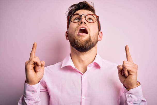 Young handsome blond man with beard and blue eyes wearing pink shirt and glasses amazed and surprised looking up and pointing with fingers and raised arms. - Photo, Image
