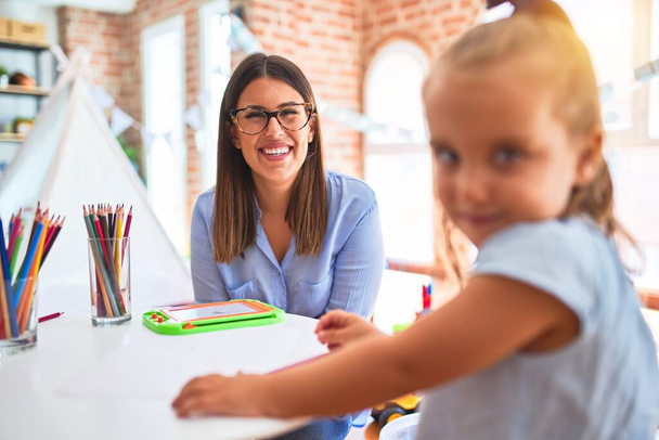 Biała dziewczynka bawiąca się i ucząca w przedszkolu z nauczycielką. Matka i córka w pokoju zabaw wokół zabawek rysunek w kolorach ołówka - Zdjęcie, obraz