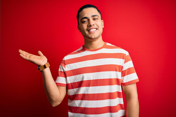 Mladý brazilský muž na sobě ležérní pruhované tričko stojící nad izolovaným červeným pozadím s veselým úsměvem prezentující a ukazující dlaní ruky při pohledu na fotoaparát. - Fotografie, Obrázek