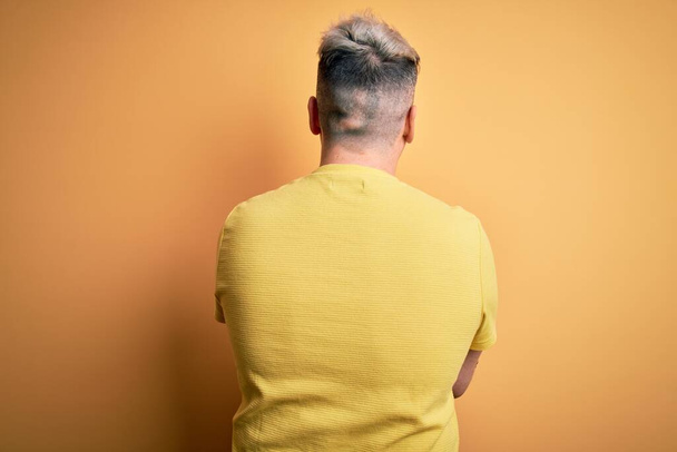 Молодий красивий сучасний чоловік у жовтій сорочці над жовтим ізольованим тлом, стоячи позаду, дивлячись з схрещеними руками
 - Фото, зображення